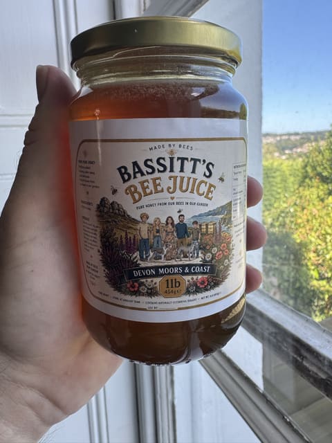 A jar of Bassitt's Bee Juice held up to the window, the honey glowing amber, with Newton Abbot's green hillside outside.
