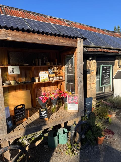 The honey cart outside Home Farm Café in the sun: a timber stall under a corrugated roof with shelves of local ceramics, honey and flowers, next to the café's stone wall and cake board.